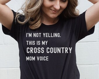 Könnte beinhalten: Schwarzes T-Shirt mit weißem Text: „I'M NOT YELLING. THIS IS MY CROSS COUNTRY MOM VOICE.“ Das Shirt ist an der Taille geknotet. Die Person trägt eine blaue Jeans. Der Hintergrund ist eine weiße Backsteinmauer.