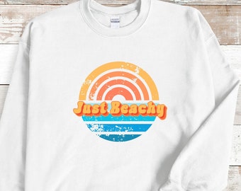 May include: White sweatshirt with a retro-style graphic of a sunset with the text "Just Beachy" in orange.