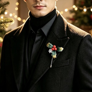 May include: A lapel pin featuring three red berries and green leaves with a small white snowflake accent attached to a long silver metal pin. The accessory is fastened to the lapel of a black wool coat worn over a black turtleneck sweater.