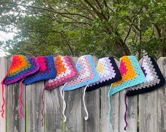 May include: A collection of colorful, crocheted head coverings, each with a unique color scheme. The head coverings are displayed on a wooden fence against a backdrop of green foliage. Each piece features a square design with a long, dangling tie.