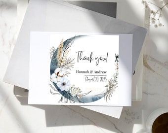 May include: A white card with a watercolor design and the words "Thank you!" in black script. The card features a floral wreath with cotton and greenery. The names "Hannah & Andrew" and the date "August 20, 2023" are also printed.