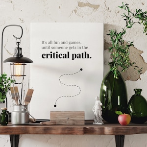 May include: A white canvas print with the text "It's all fun and games, until someone gets in the critical path." with a dotted line path and two black dots. The print is on a wooden shelf with a green plant, a lamp, a vase with a green plant, a green vase, a red apple, and a small statue.