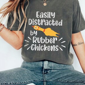 May include: Dark grey t-shirt featuring the text "Easily Distracted by Rubber Chickens" and a yellow rubber chicken illustration. The top is styled with blue denim jeans.
