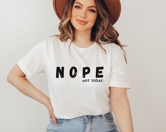 May include: White t-shirt with the text "NOPE NOT TODAY" in black, distressed font. The shirt is paired with blue jeans and a brown hat. The overall style is casual and relaxed.