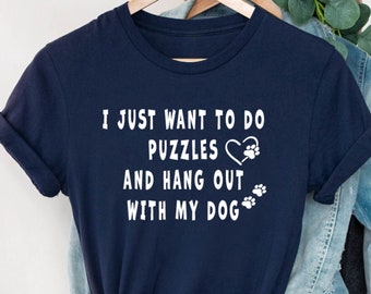 Może przedstawiać: Granatowa koszulka z białym napisem: "I JUST WANT TO DO PUZZLES AND HANG OUT WITH MY DOG." Zawiera serce, odcisk łapy i grafiki odcisków łap. Koszulka wisi na drewnianym wieszaku.
