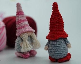 May include: Two crocheted gnomes, one with a pink and white striped hat and the other with a red hat. Both gnomes have gray bodies and white beards.