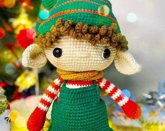 May include: A crocheted elf doll with a green hat, brown hair, and a green and white striped outfit. The elf is sitting on a white surface with string lights.