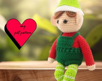 May include: A crocheted elf doll with a green hat, red shirt, green pants, and white and green striped leggings. The elf is sitting on a wooden surface. The text "eng pdf pattern" is in a red heart shape.