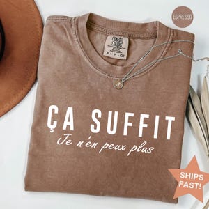 May include: An espresso-colored short-sleeve t-shirt featuring the white text "ÇA SUFFIT" in a bold sans-serif font, followed by the white cursive text "Je n'en peux plus". The shirt has a crew neckline and a Comfort Colors brand label.