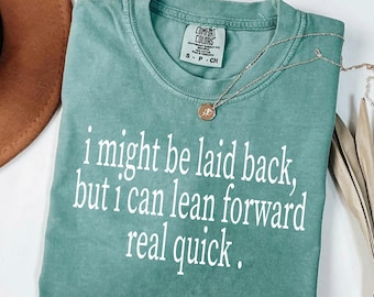 May include: A seafoam green t-shirt with the text "i might be laid back, but i can lean forward real quick" in white. A silver necklace with a pendant is on the shirt. The shirt is folded on a white surface.
