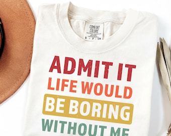 May include: Cream-colored t-shirt with the text "ADMIT IT LIFE WOULD BE BORING WITHOUT ME" in red, orange, yellow, and green. The shirt is folded and has a Comfort Colors tag.