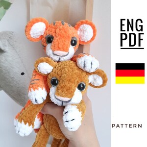 May include: Two crocheted tiger plushies, one orange and one brown, with white accents. The orange tiger is holding the brown tiger in its paws. The image includes the text "ENG PDF PATTERN".