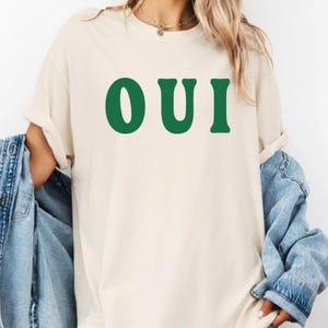 May include: A beige short-sleeved t-shirt featuring the word OUI printed in large, bold, dark green capital letters across the chest. The shirt has a crew neckline and a relaxed fit, paired with a blue denim jacket draped over the shoulders.