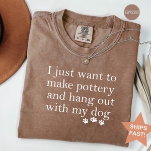 May include: Espresso-colored t-shirt with the text "I just want to make pottery and hang out with my dog" in white. The shirt has a crew neck and a Comfort Colors tag. A silver necklace with a pendant is visible.