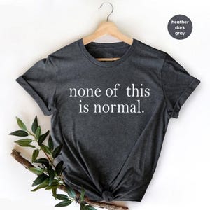 May include: Dark heather grey t-shirt with the white text "none of this is normal". The t-shirt is hanging on a wooden hanger against a white backdrop. A sprig of greenery is placed in the lower left corner of the image.