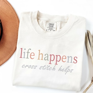 May include: Cream-coloured t-shirt with the text "life happens cross stitch helps" in colourful lettering. The shirt has a crew neck and short sleeves. The brand tag "Comfort Colors" is visible at the neckline.