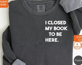 May include: Dark gray long-sleeve sweatshirt with the text "I CLOSED MY BOOK TO BE HERE" in white. The sweatshirt has a crew neck and a soft, fleece-lined interior. The Comfort Colors brand tag is visible.
