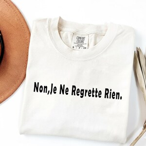 May include: A cream-coloured short-sleeve t-shirt with a crew neckline. The shirt features the black printed text "Non, Je Ne Regrette Rien." across the chest. A white interior neck label displays the brand name Comfort Colors and size S.