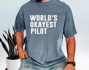 May include: A blue-grey t-shirt with the text "WORLD'S OKAYEST PILOT" in white block letters. The shirt is a casual style, likely made of cotton, and is being worn by a person. The overall aesthetic is relaxed and comfortable.