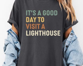 May include: Dark grey t-shirt with the text "IT'S A GOOD DAY TO VISIT A LIGHTHOUSE" in teal, yellow, orange, and off-white. The shirt is a casual style, suitable for everyday wear.