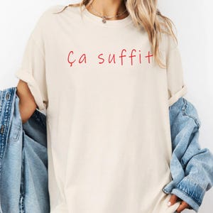 May include: A beige short-sleeve t-shirt featuring the red text "Ça suffit" printed across the chest. The shirt has a crew neckline and is worn with a blue denim jacket draped over the shoulders, showing the sleeves and front panels of the jacket.