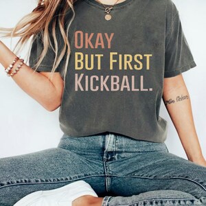 Peut inclure: T-shirt gris foncé avec l'inscription "Okay But First Kickball" en rose et jaune. Un haut décontracté et confortable pour un usage quotidien.