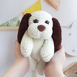 Può includere: Un peluche a forma di cane all'uncinetto fatto a mano, color crema e marrone. Il cane ha orecchie marroni flosce, un naso marrone e occhi neri. Il peluche è morbido e coccoloso, perfetto per i bambini o come oggetto decorativo.