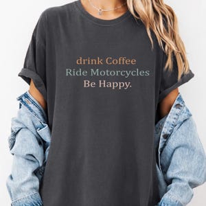 以下が含まれることがあります： 「drink Coffee Ride Motorcycles Be Happy」の文字が入った濃いグレーのTシャツ。テキストはオレンジ、ティール、ピンクです。シャツは袖をまくり、デニムジャケットを着ています。