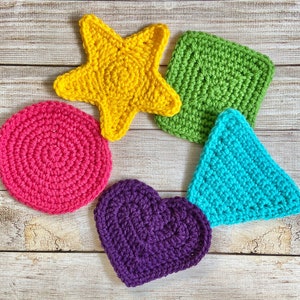 Op de afbeelding: Een verzameling kleurrijke, gehaakte vormen: een gele zeester, een groene vierkant, een roze cirkel, een turquoise driehoek en een paars hart. Gerangschikt op een licht houten oppervlak.