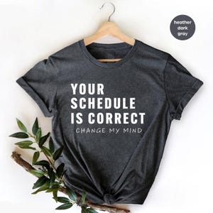 Op de afbeelding: Donkergrijze heather t-shirt met de witte tekst "YOUR SCHEDULE IS CORRECT CHANGE MY MIND". Het t-shirt hangt aan een houten hanger.