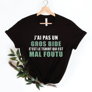 Peut inclure: T-shirt noir avec le texte français "J'AI PAS UN GROS BIDE C'EST LE TSHIRT QUI EST MAL FOUTU" en blanc et vert. Le t-shirt est suspendu à un cintre en bois sur fond blanc, avec des feuilles vertes et une branche en bas à gauche.