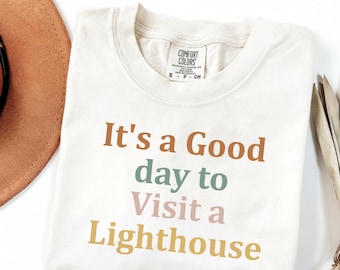 May include: Cream-coloured t-shirt with the phrase "It's a Good day to Visit a Lighthouse" in brown, teal, pink, and yellow lettering. The shirt is folded and has a Comfort Colors tag. A brown hat and decorative item are partially visible.
