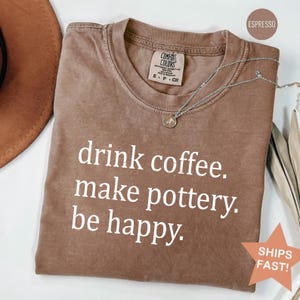 Może przedstawiać: Koszulka w kolorze espresso z napisem "drink coffee. make pottery. be happy." Srebrny naszyjnik z wisiorkiem spoczywa na koszulce. Koszulka ma metkę Comfort Colors i naklejkę "SHIPS FAST!"