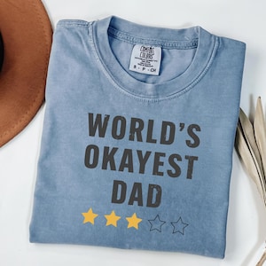 Puede incluir: Una camiseta azul con el texto "WORLD'S OKAYEST DAD" en gris oscuro. Debajo, tres estrellas amarillas y dos estrellas delineadas. La camiseta tiene cuello redondo y una pequeña etiqueta en el pecho.