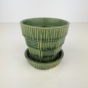 May include: A sage green ceramic plant pot with a matching saucer, featuring a textured basket-weave pattern of vertical and horizontal lines. The pot has a wide, flared rim and a tapered body, with a dark green glaze accentuating the relief details.