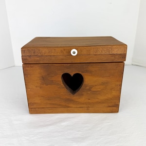 Pode incluir: Uma caixa de madeira com um recorte em forma de coração na frente. A caixa é feita de madeira de tons quentes e tem uma tampa articulada presa por um pequeno botão branco. Ideal para armazenamento.