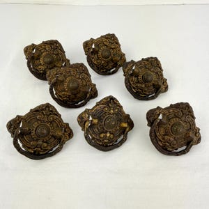 May include: Seven antique bronze drawer pulls with ornate, detailed designs. Each pull features a central circular element surrounded by scrolling patterns and a curved handle. The pulls are aged with a dark, weathered finish, suggesting vintage craftsmanship.