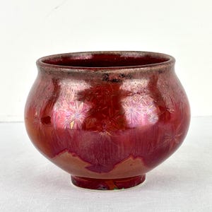 May include: A rounded ceramic bowl featuring a deep red crystalline glaze with iridescent starburst patterns. The rim has a dark, metallic-toned edge, and the base is narrow, transitioning into a wide, bulbous body with a glossy, reflective finish.