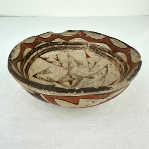 May include: A shallow ceramic bowl featuring a cream-colored base with hand-painted geometric patterns in dark brown and terracotta red. The rim displays a repeating zigzag motif, while the interior contains angular, abstract line designs.