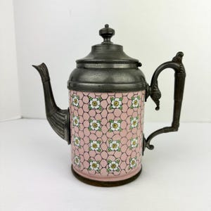 May include: Antique metal teapot with a pink floral design. The teapot features a dark metal lid, handle, and spout. The body of the teapot is decorated with a pink background and a white and yellow floral pattern. The teapot has a height of approximately 8 inches.