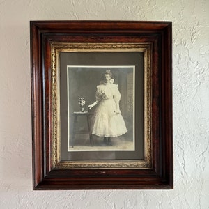 May include: A framed black and white photograph of a woman in a white dress, standing next to a small table with flowers. The frame is dark brown with gold accents, mounted on a textured white wall. The photograph is vintage.