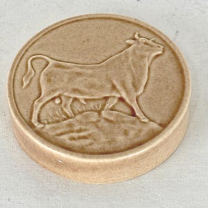 Peut inclure: Un disque en céramique rond de couleur brun clair présentant un relief en saillie représentant un taureau debout sur un sol texturé. La surface possède une finition lisse et brillante avec des variations subtiles de la teinte beige.