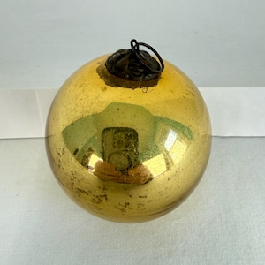 May include: A vintage, round Christmas bauble with a gold finish. The bauble has a dark metal cap and a loop for hanging. The surface shows signs of age and wear, adding to its antique charm. A reflection is visible on the surface.