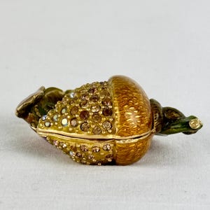 May include: A decorative, hinged trinket box shaped like a golden acorn. The acorn's cap is adorned with sparkling crystals, while the body features a textured, painted finish. The stem is green with gold accents. The box is open, revealing the interior.