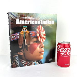 Może przedstawiać: Twarda oprawa książki zatytułowana "The World of the American Indian" z portretem Indianina na okładce. Osoba nosi opaskę z koralików z piórami i czerwoną farbą do twarzy. Czerwona puszka Coca-Coli znajduje się obok książki.