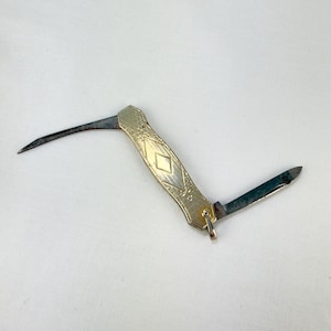 May include: A small folding pocket knife with a gold-toned metal handle featuring an engraved diamond pattern. The knife has two thin, dark, weathered steel blades extended in opposite directions and a small metal loop attached to one end.