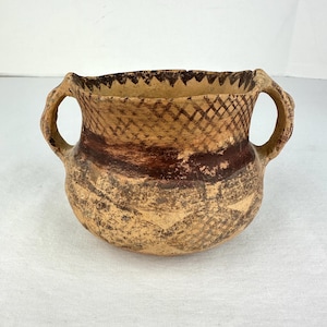May include: A tan ceramic pot with two side handles, featuring a dark brown painted cross-hatch pattern around the upper rim and a jagged zigzag design along the top edge. The lower body displays a faded, weathered geometric pattern in brown.