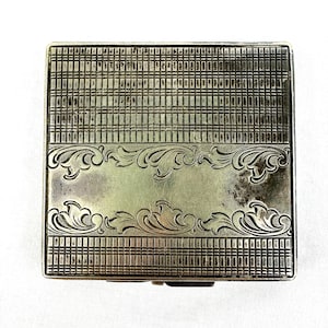 May include: A silver-toned, rectangular compact with a detailed, engraved design. The top features a grid-like pattern above and below a central panel with ornate, floral scrollwork. The compact is closed and appears to be vintage.