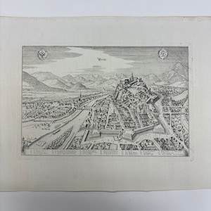 May include: An antique black and white print of Graz, Austria, featuring a detailed bird's-eye view of the city, including buildings, a river, and surrounding mountains. The print includes the word "Graz" at the top and several numbered labels.