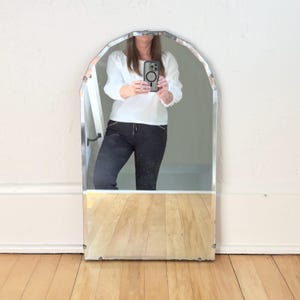 May include: An arched wall mirror with a frameless design and beveled edges. The mirror features a rounded top section that transitions into straight vertical sides. The reflective surface shows a clear view of a person wearing a white top and black pants.